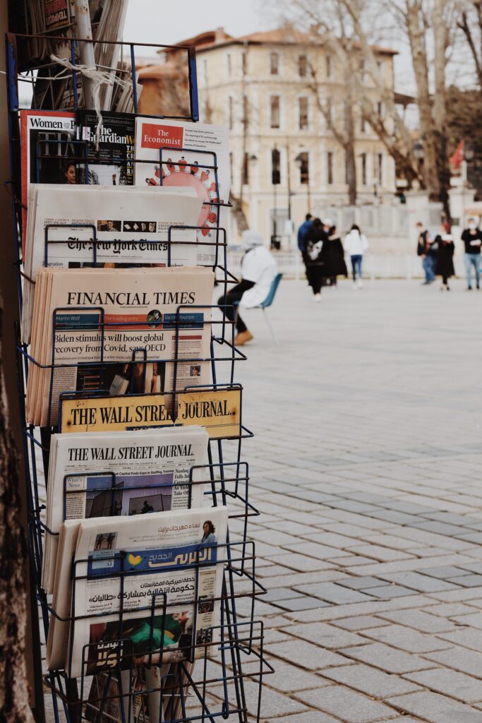 A rack of newspapers for sale in a brick plaza, with people walking by in the background.